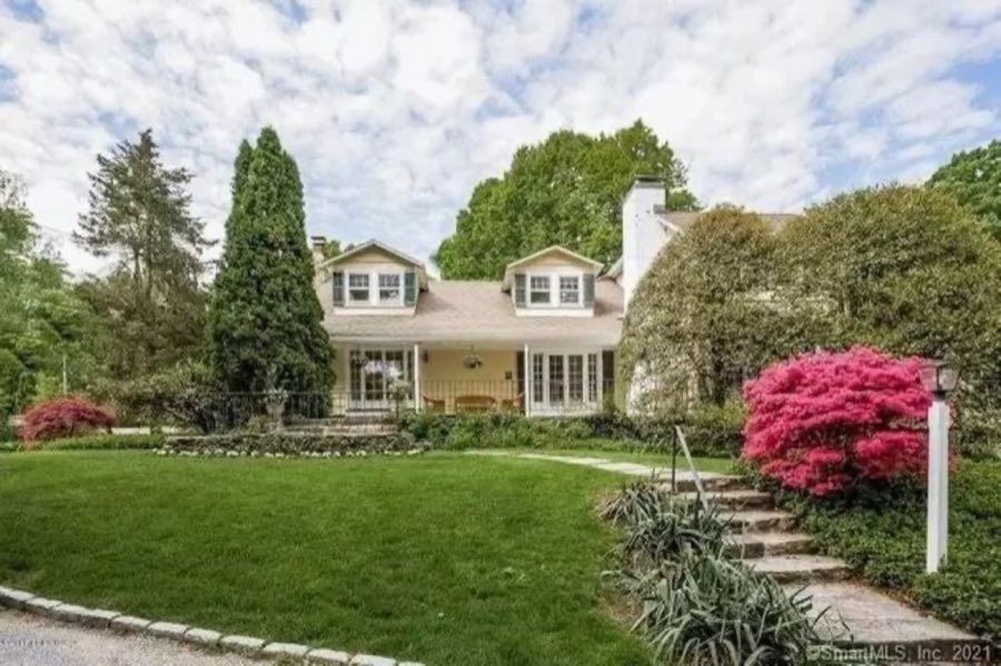 Cream clapboard house with dormer windows and a columned porch, framed by a tall conifer and flowering azaleas across a mown lawn.