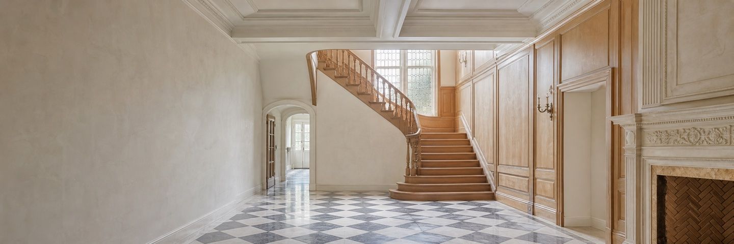 Illustration — An empty hall with a curved staircase in pale timber, its turned balusters rising past a tall leaded window. The floor is grey and white marble laid as a chequerboard. Panelled walls and a carved stone chimneypiece stand to the right, an arched doorway beyond.