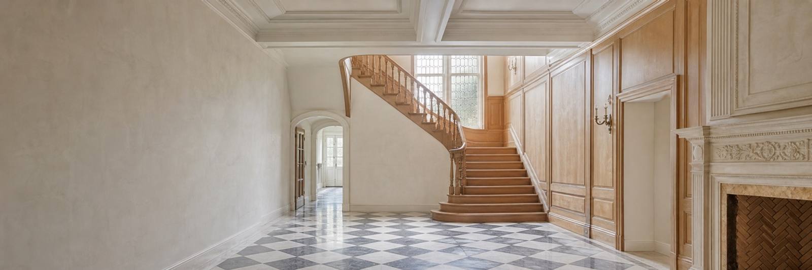 Illustration — An empty hall with a curved staircase in pale timber, its turned balusters rising past a tall leaded window. The floor is grey and white marble laid as a chequerboard. Panelled walls and a carved stone chimneypiece stand to the right, an arched doorway beyond.