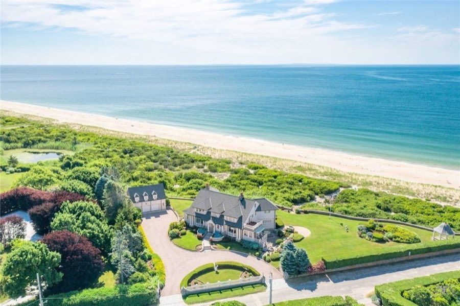 Aerial view of a shingled oceanfront house and detached garage on a lawn behind dunes, with a curved gravel drive and a long sand beach meeting open ocean.