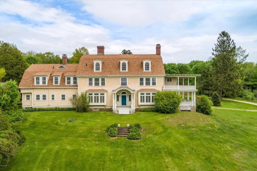 Pale yellow clapboard colonial with a shingled gambrel roof, arched dormer windows, two brick chimneys and a covered side porch, standing on a broad lawn.