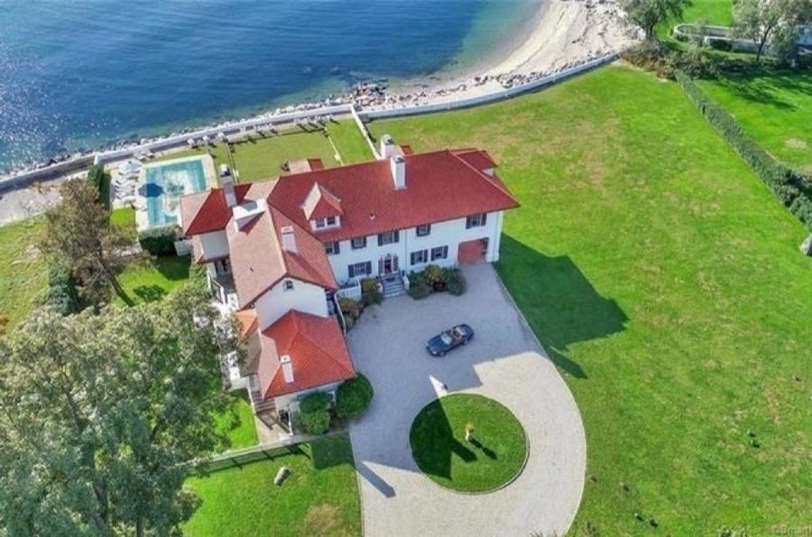 Aerial view of a white waterfront house with a red tile roof, a curved gravel drive and a pool terrace above a seawall and pebble beach.