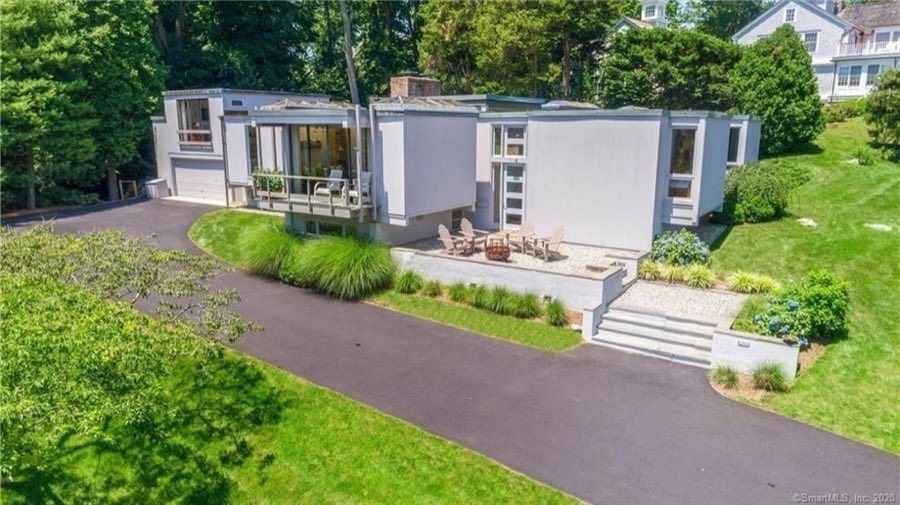 Mid-century modern house in white board-and-batten with flat rooflines, clerestory windows and a deck, above a gravel fire-pit terrace and a curving drive.