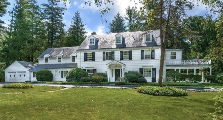 White clapboard colonial with black shutters, dormer windows, a pedimented entry and an attached garage wing, across a broad mown lawn.