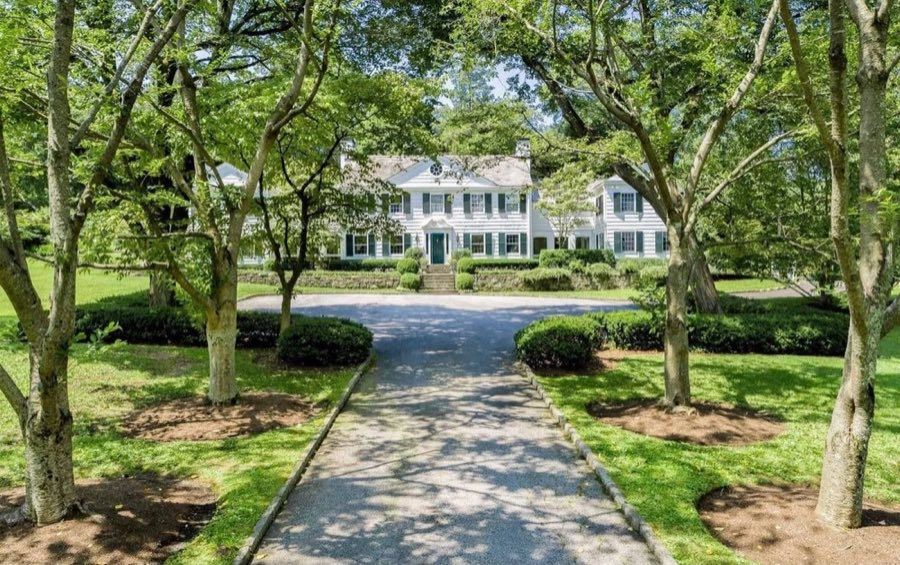 White clapboard colonial with dark shutters and a pedimented entry, seen down a long tree-lined drive between clipped hedges and mown lawn.