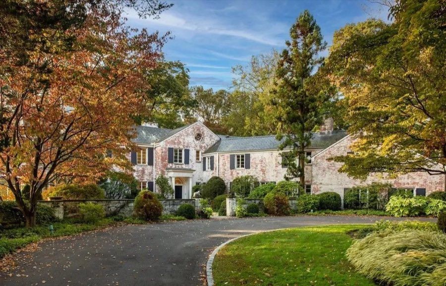 Whitewashed brick house with dark shutters, a pedimented entry and a slate roof, seen across a curving drive framed by autumn maples.