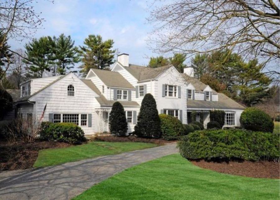 White shingled colonial with grey shutters, gambrel dormers and clipped topiary along a curving drive, under bare winter branches.