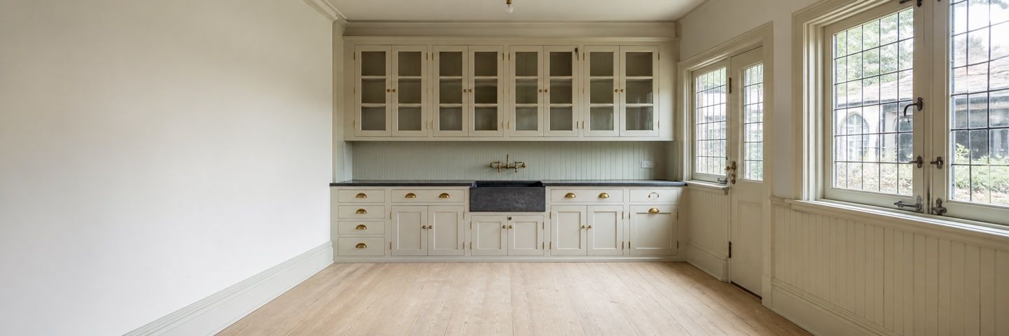 Illustration — An empty butler's pantry. Cream glazed-front cupboards with brass cup handles run above a dark stone worktop and a deep farmhouse sink with a brass bridge tap, over beadboard panelling and plain cupboard doors. A glazed door and leaded windows open to the right.