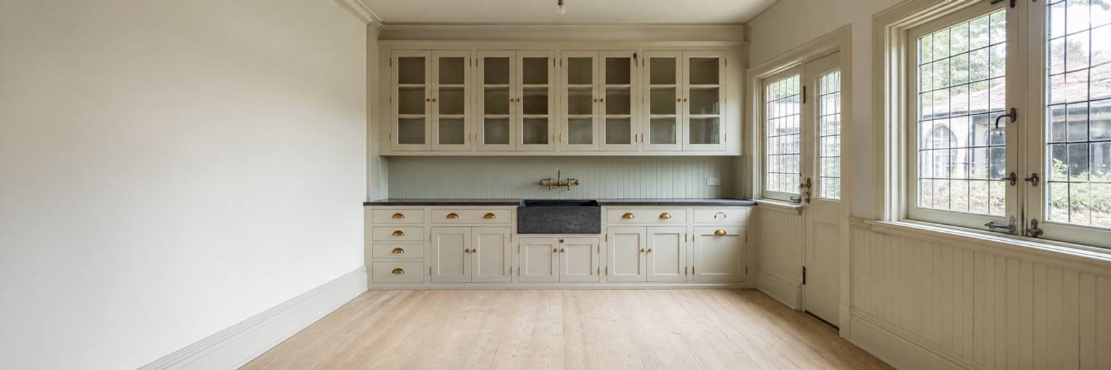 Illustration — An empty butler's pantry. Cream glazed-front cupboards with brass cup handles run above a dark stone worktop and a deep farmhouse sink with a brass bridge tap, over beadboard panelling and plain cupboard doors. A glazed door and leaded windows open to the right.
