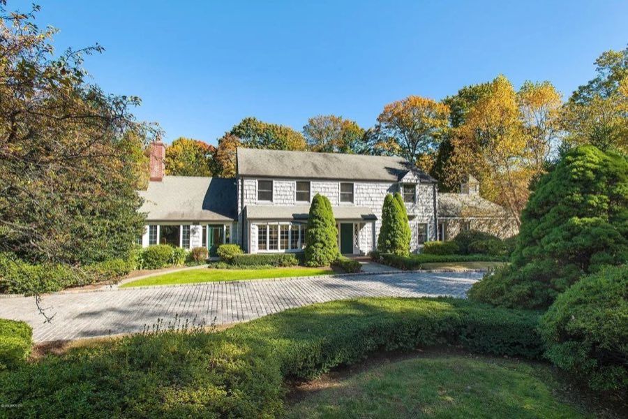 Grey shingled colonial with a green front door, bay windows and a brick chimney, behind a wide cobbled forecourt and clipped box hedging in autumn.