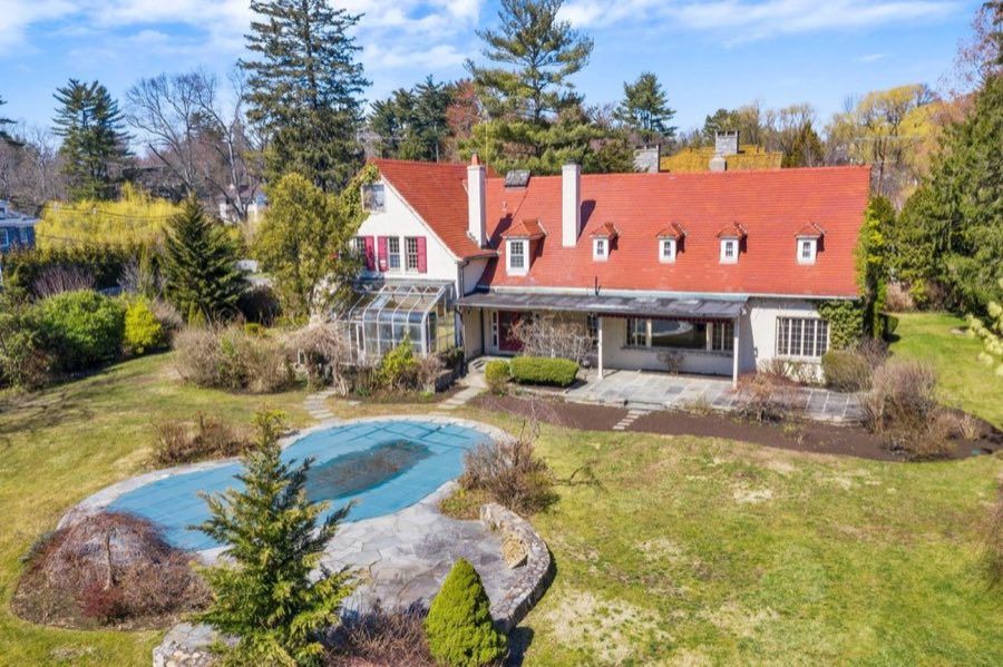 Aerial view of a stucco house with a steep red tile roof and dormers, a glass conservatory, a covered terrace and a kidney-shaped covered pool on the lawn.