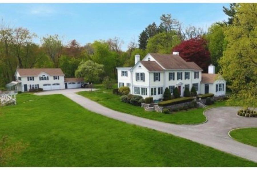 Aerial view of a white colonial with black shutters and a detached garage barn, joined by a curving drive across a wide lawn.