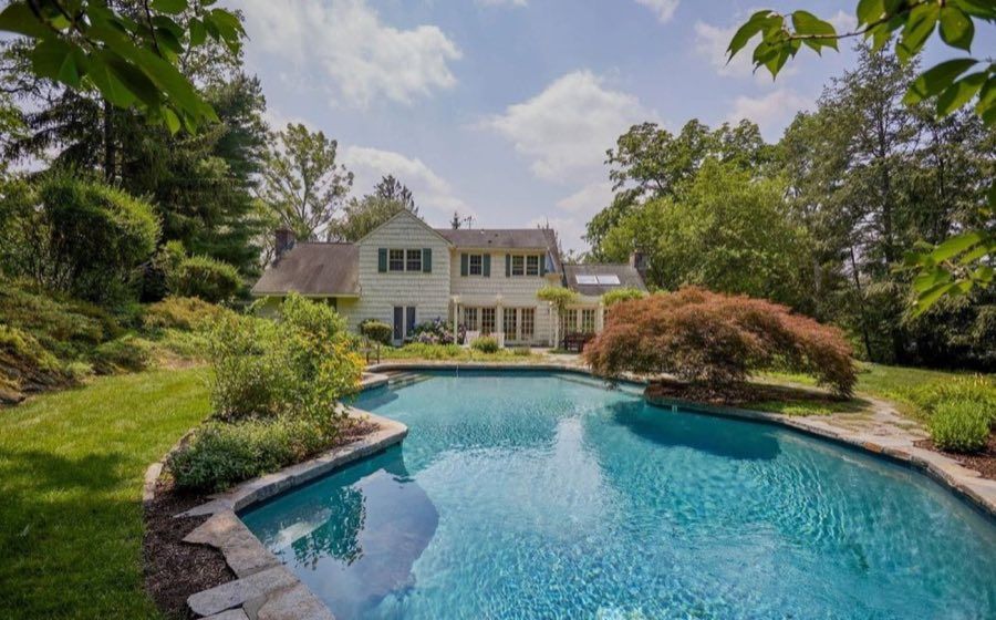 Cream clapboard colonial with green shutters seen across a curved stone-edged swimming pool, with a red Japanese maple beside the terrace.