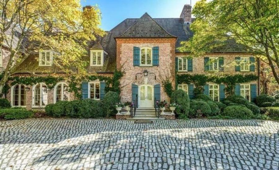 Brick country house with a steep slate roof, dormer windows, teal shutters and climbing ivy, behind a fan-patterned cobblestone motor court and clipped hedges in autumn light.