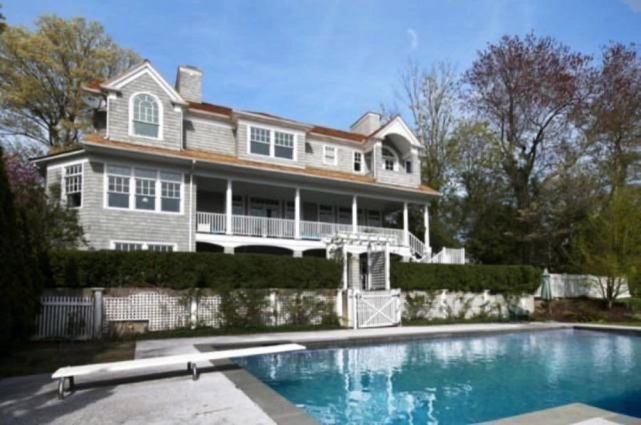 Grey shingled house with a deep columned veranda, an arched dormer and gambrel roof, above a clipped hedge and a swimming pool with a diving board.