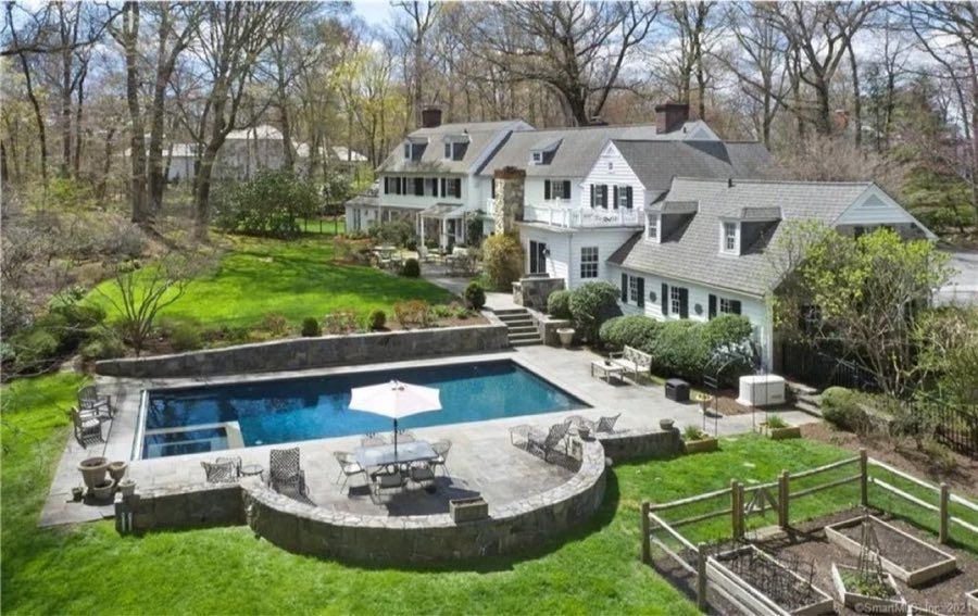 Aerial view of a white clapboard colonial with dormers and a stone chimney, above a terraced bluestone pool terrace with a rectangular pool, umbrella table and raised vegetable beds.
