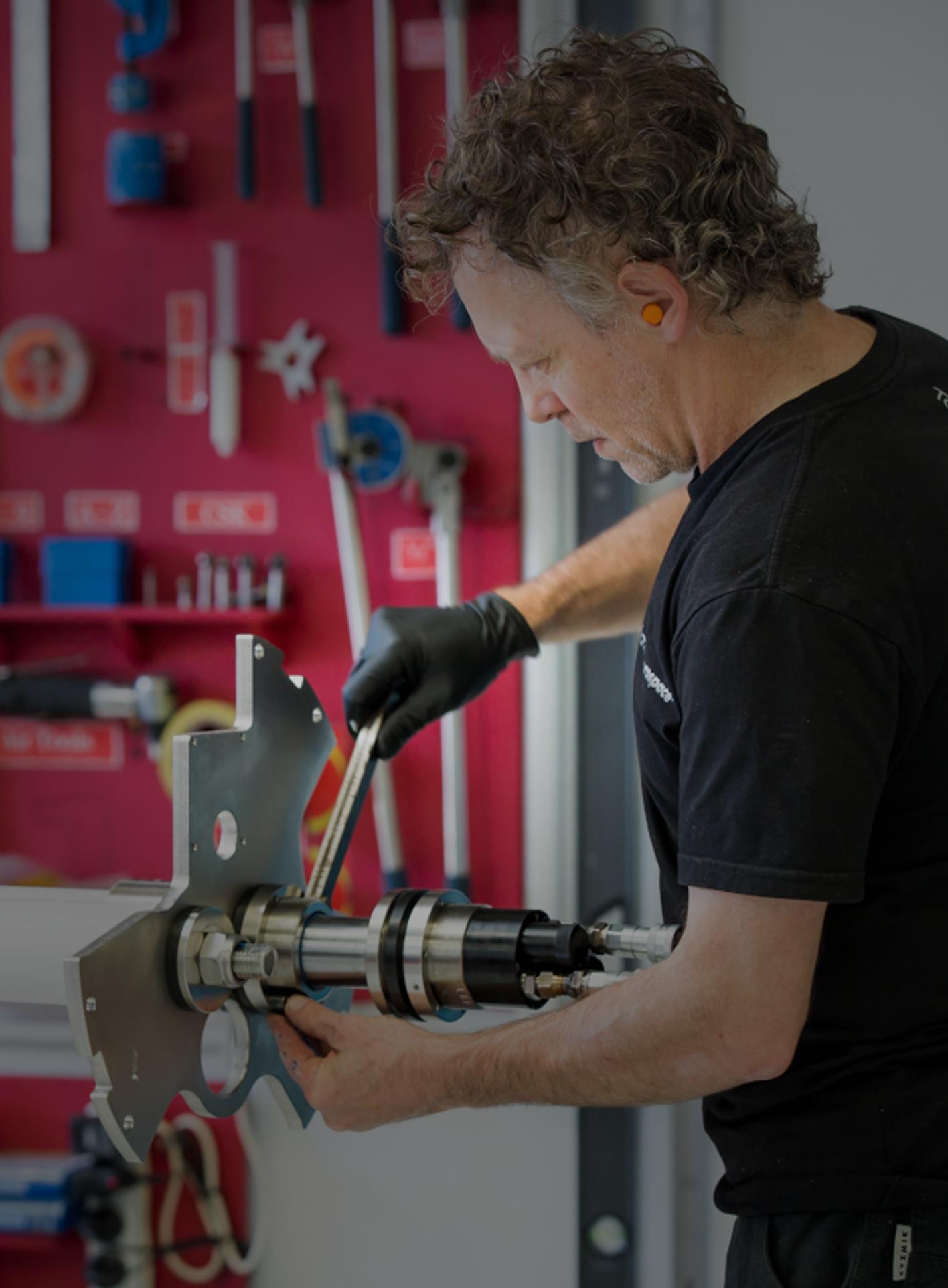 A person works with machinery in a workshop, wearing gloves and ear protection. Behind them, red tool wall organized with various tools. Focused mood.