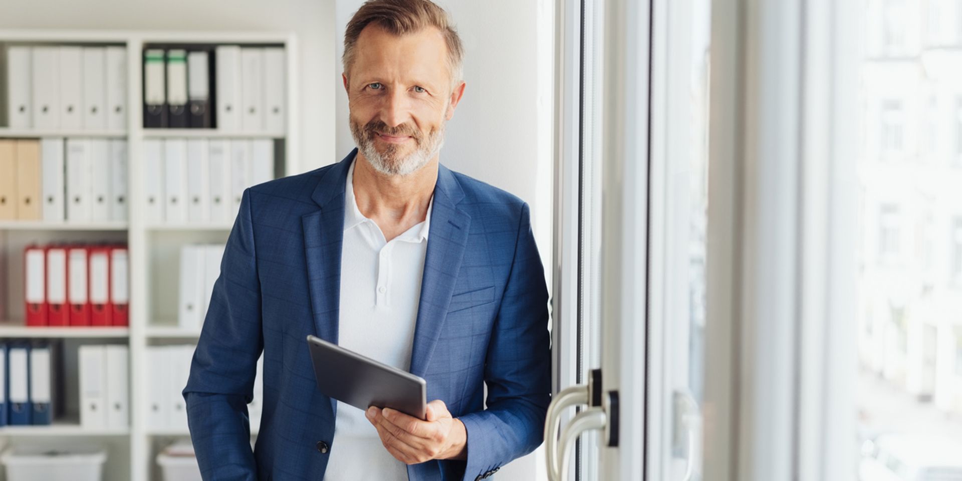 Smart relaxed confident senior businessman holding a tablet standing leaning against a glass door at the office smiling at camera