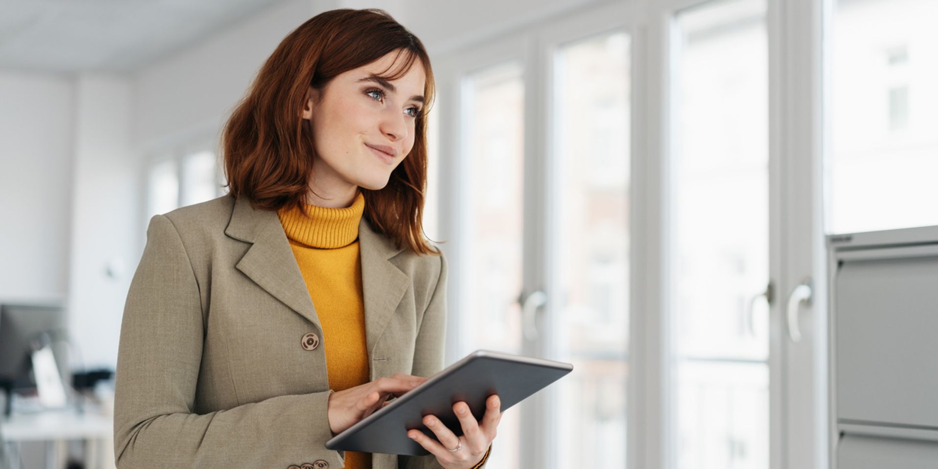 Stylish young businesswoman using a handheld tablet pc looking away with a quiet smile in a close up low angle office portrait with copy space