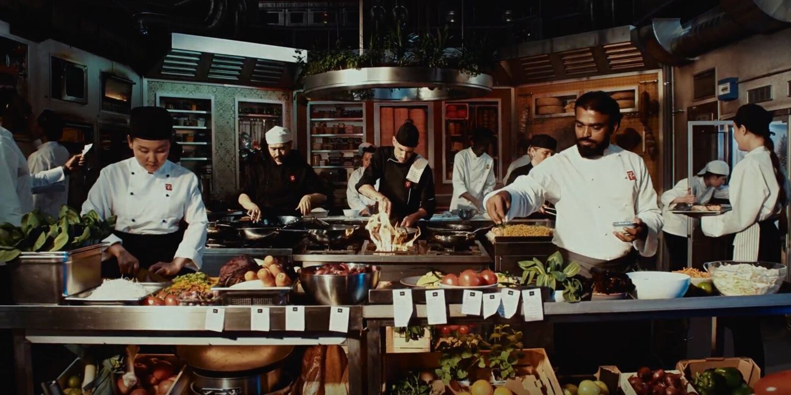 Chefs in a bustling kitchen are focused on preparing dishes with fresh ingredients. The atmosphere is lively and collaborative, showcasing culinary creativity.