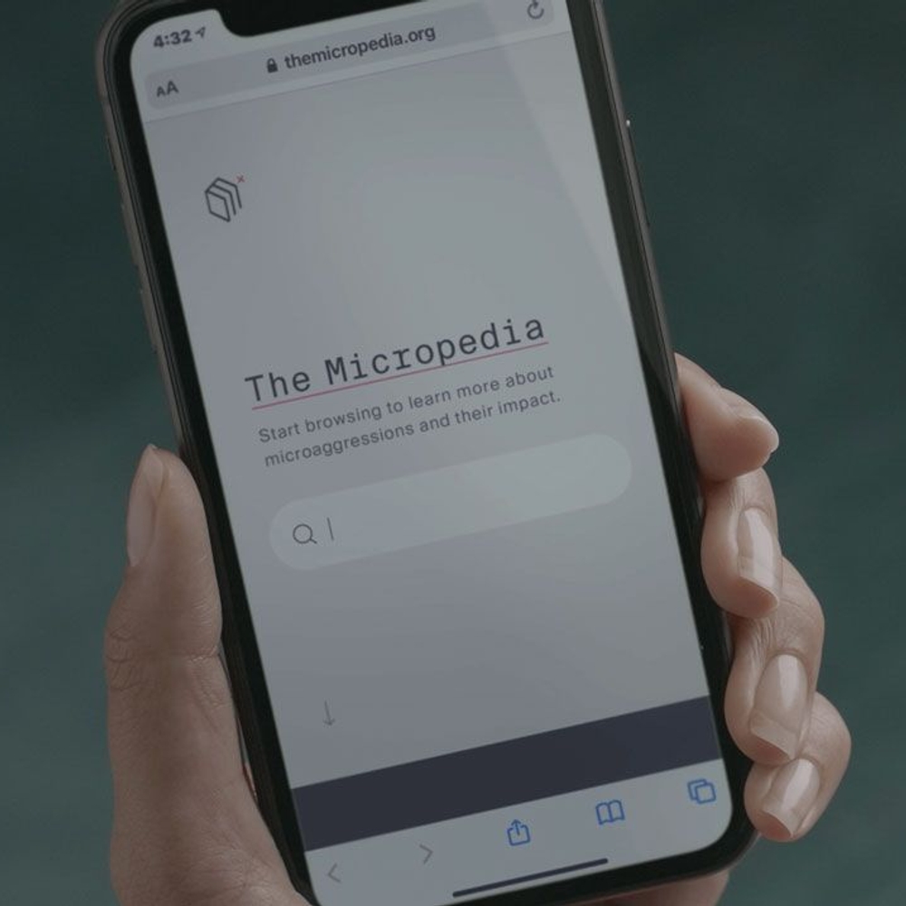 A hand holding a phone displaying the Micropedia of Microaggression website.