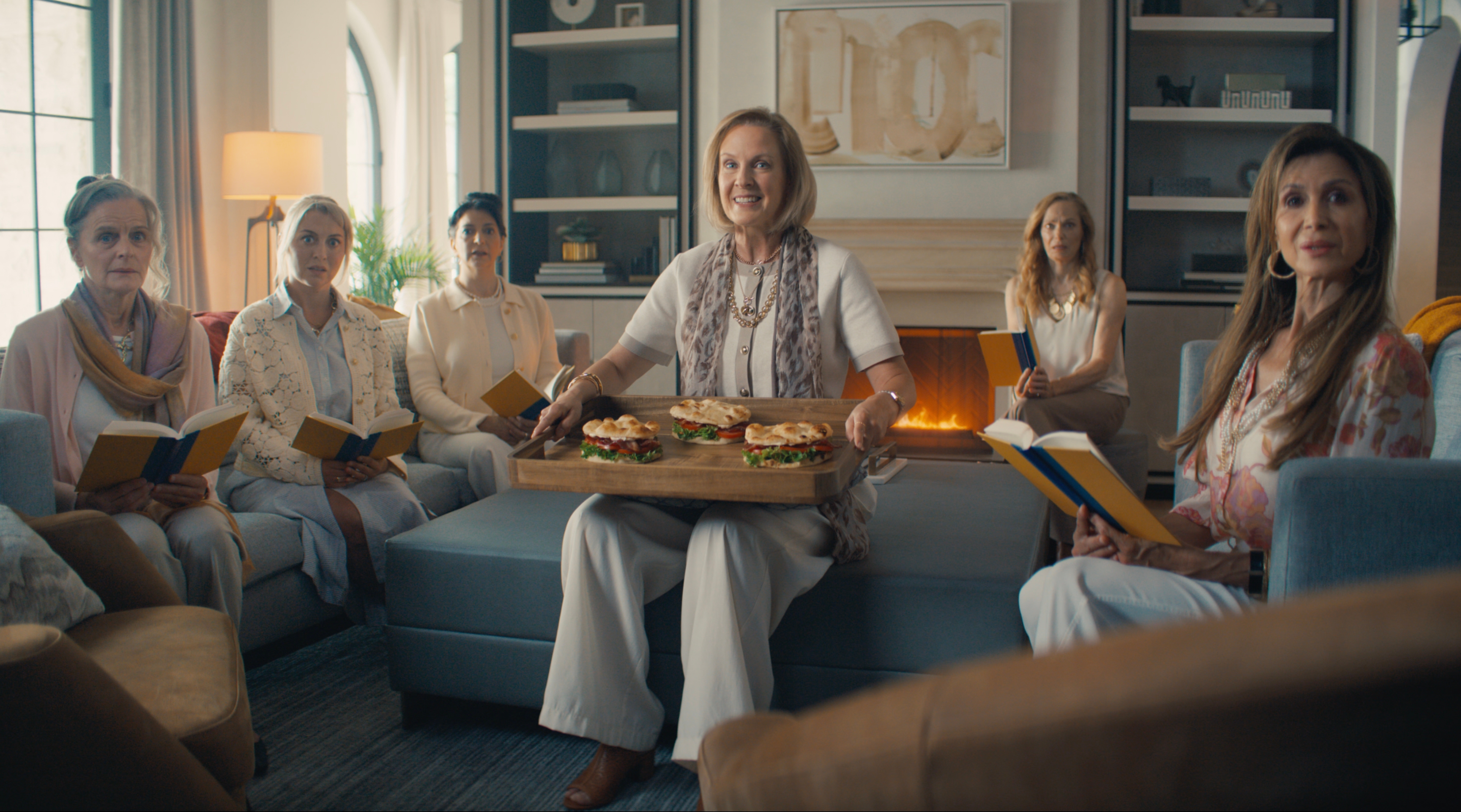 Seven women sit in a cozy living room, reading books.