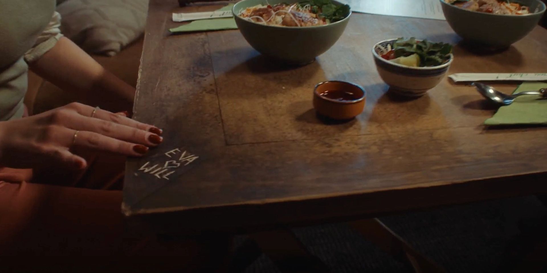 A wooden table with carved initials “Eva ♥ Will.” Three bowls of salad, two green napkins, and cutlery are neatly arranged, creating a cozy, intimate dining scene.