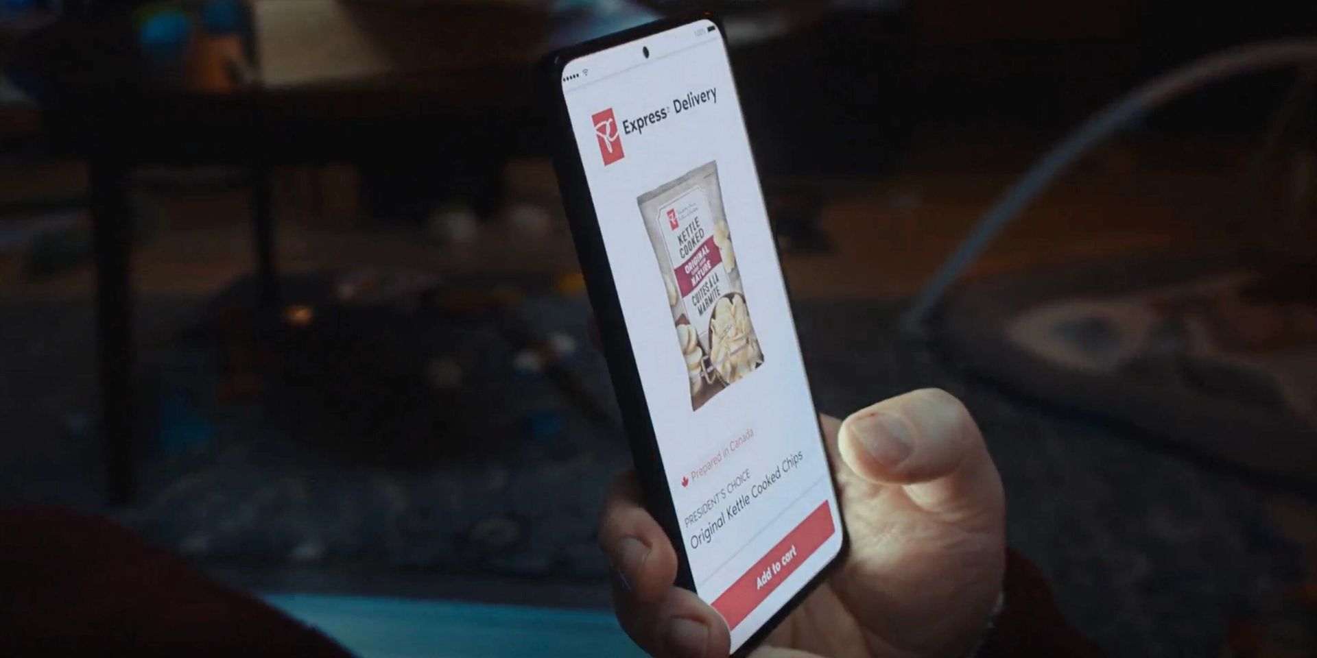A person is holding a smartphone displaying a grocery delivery app, showing a bag of kettle-cooked chips with an “Add to cart” button in a dimly lit room.