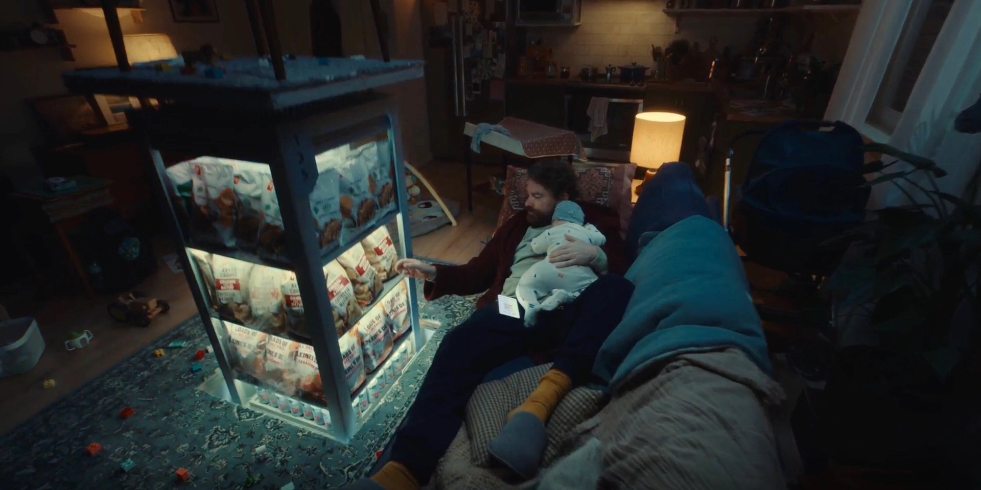 A man with a baby on his lap sits on a couch in a dimly lit living room, staring at an illuminated chicken rotisserie stand. The room is cluttered with toys, and the atmosphere feels cozy and whimsical.