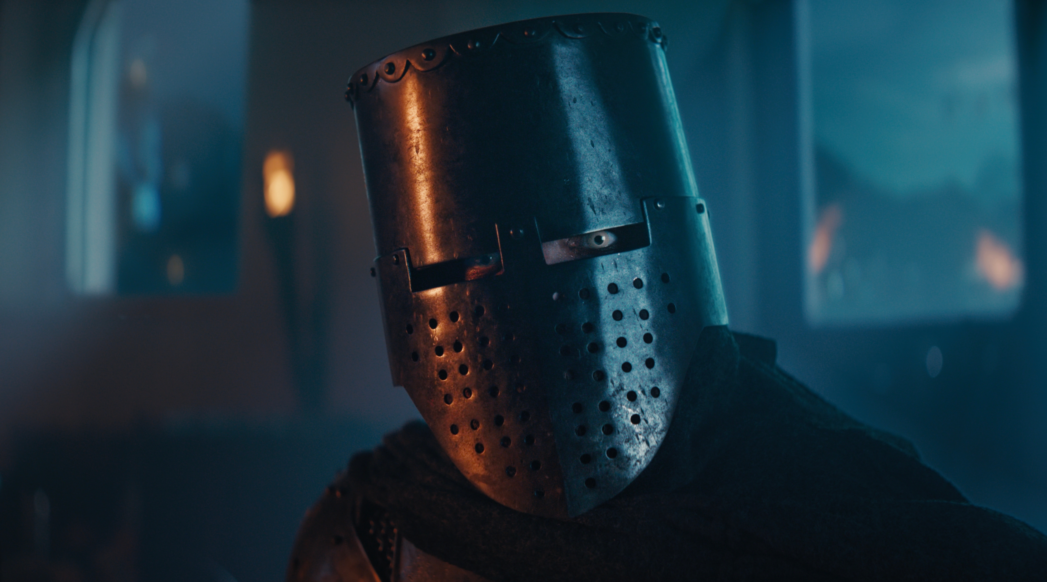 A knight in a worn, perforated helmet gazes intently in a dimly lit room.