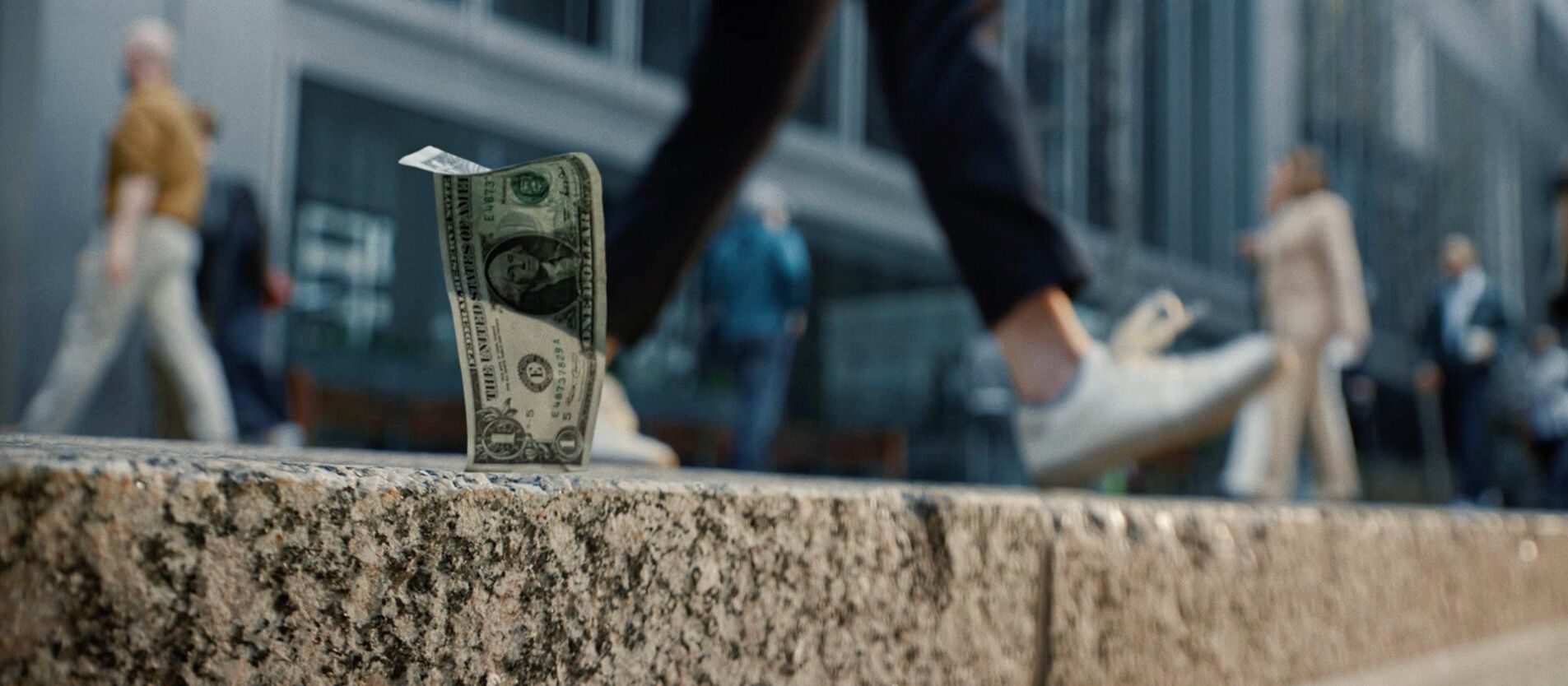 A lone dollar bill stands upright on a rain-soaked street, reflecting city lights in puddles; blurred nighttime surroundings create a moody tone.