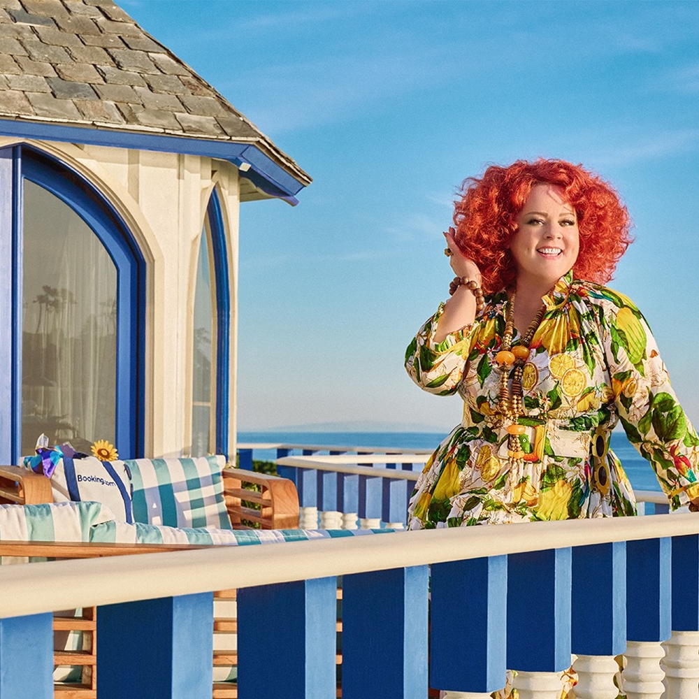 Melissa McCarthy stands on a balcony overlooking a stunning ocean view, smiling as she touches her hair.