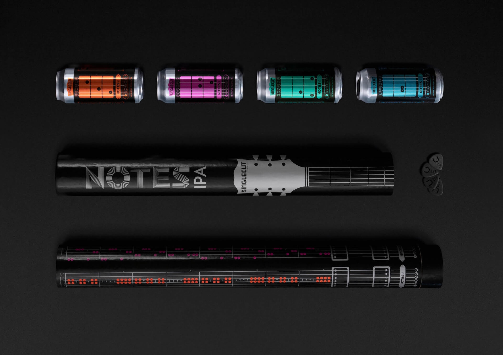 A picture featuring three Notes IPA cans with rolls of campaign packaging with guitar playing tips.