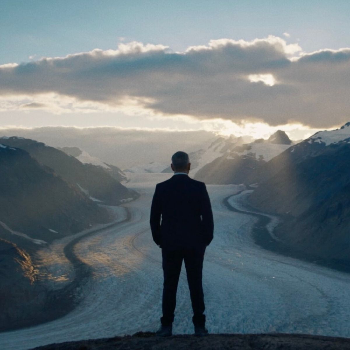 A person in a suit stands on a rock, overlooking a vast glacier flanked by mountains under a partly cloudy sky