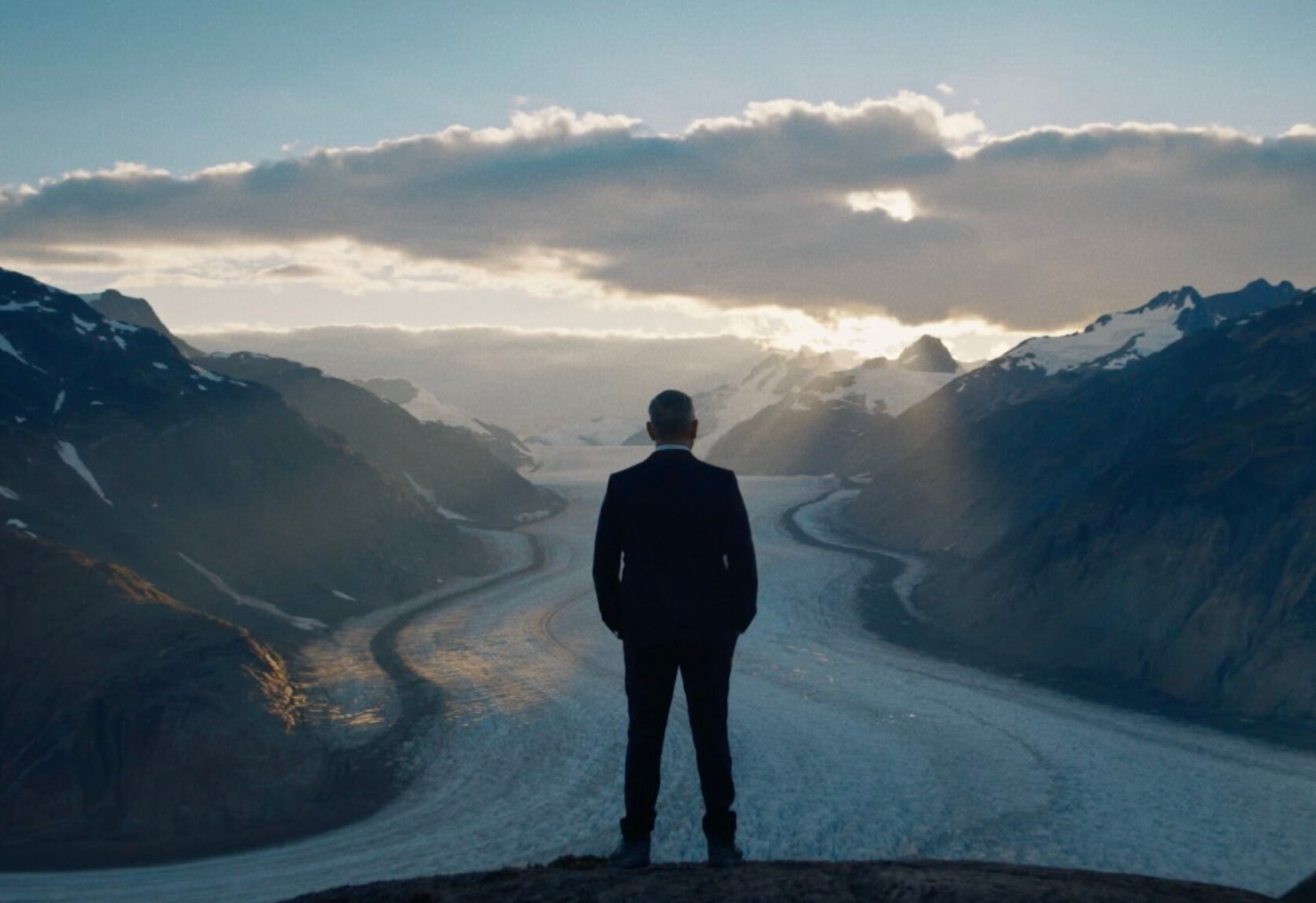 A person in a suit stands on a rock, overlooking a vast glacier flanked by mountains under a partly cloudy sky