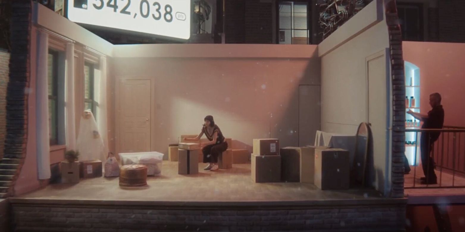 A young woman sits on a box in a dimly lit, partially furnished room with scattered boxes. A man stands on a balcony with a blue light. The mood is reflective.