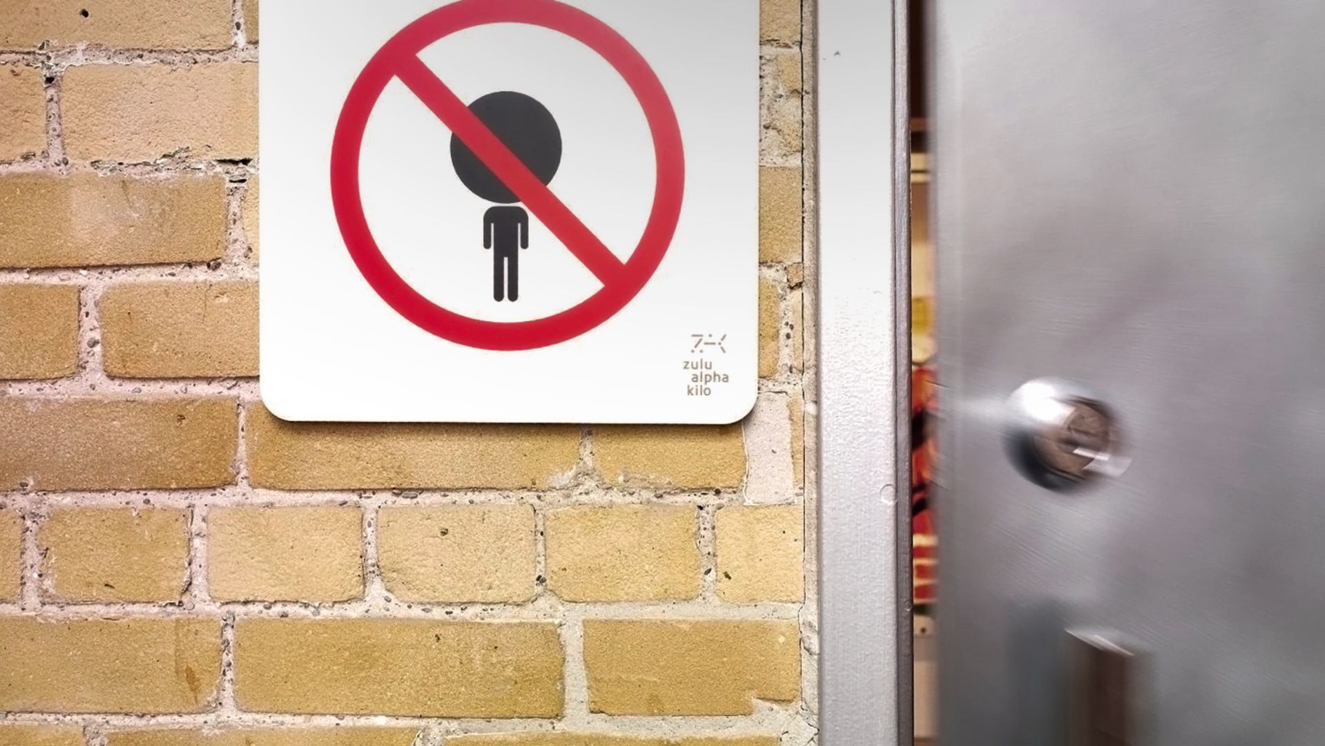 sign on a brick wall shows a stick figure with a large head crossed out, indicating “No Ego.” A partially open metal door is beside it.
