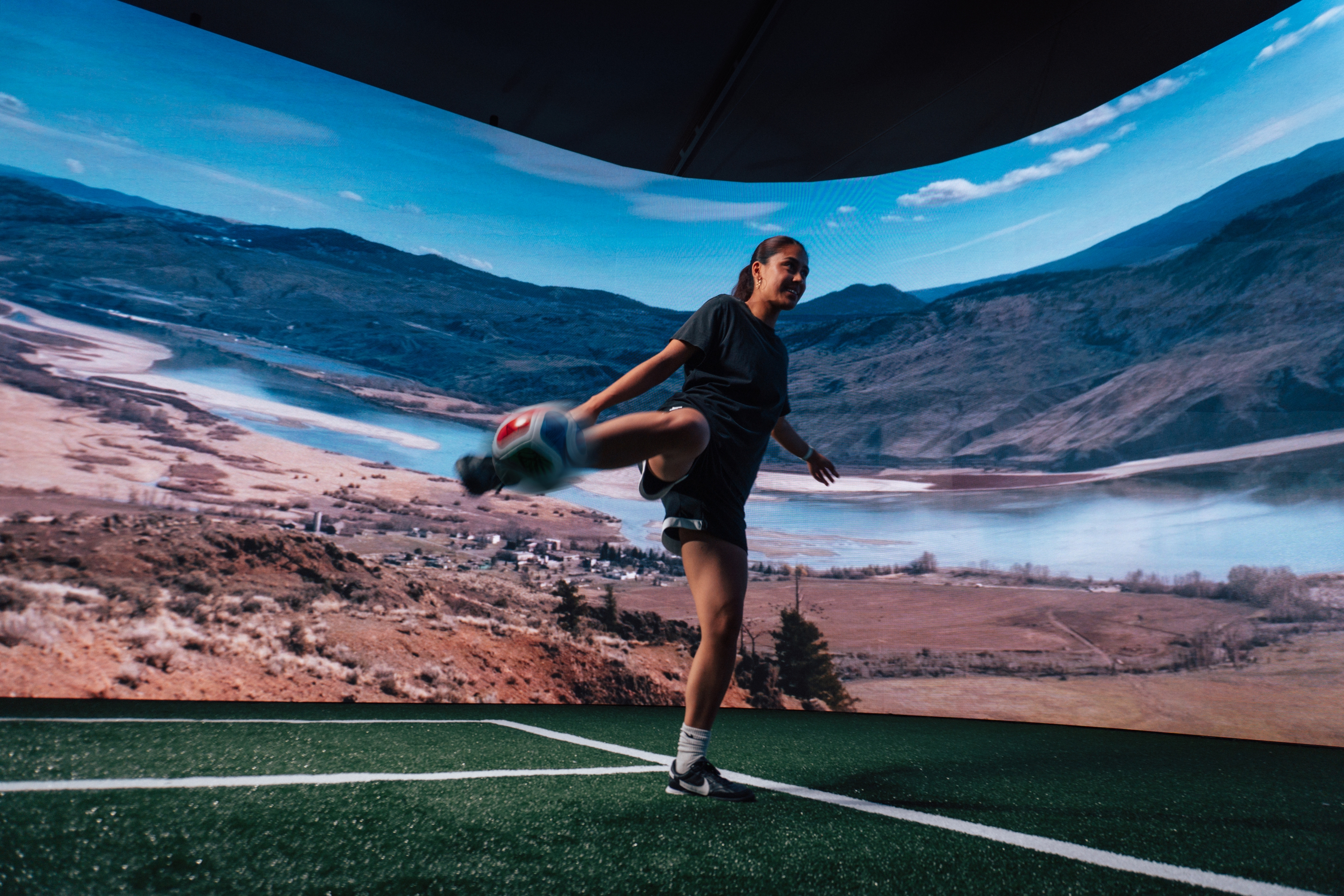 A soccer player in black balances mid-kick on indoor turf before a curved mountain landscape screen, creating an energetic, adventurous scene.