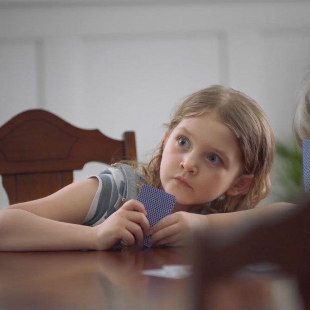 A young girl sitting at the table, holding cards, looking very surprised.