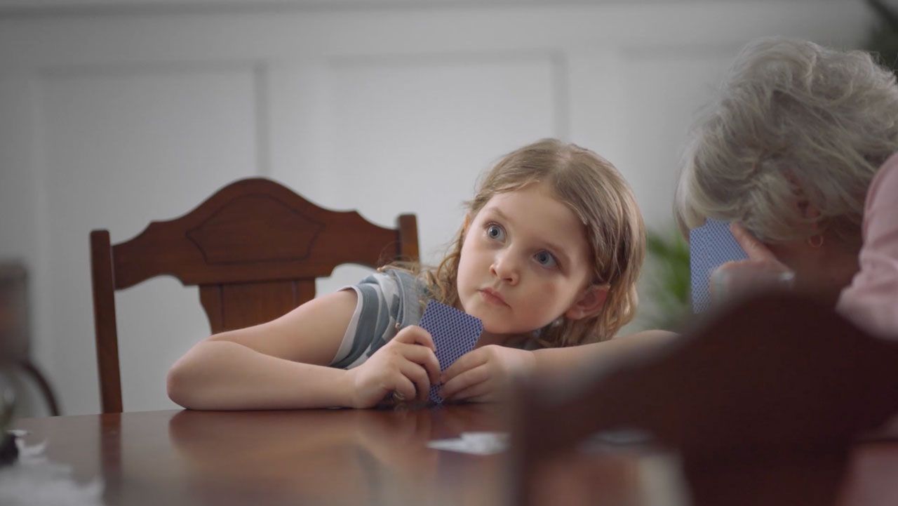 A young girl sitting at the table, holding cards, looking very surprised.