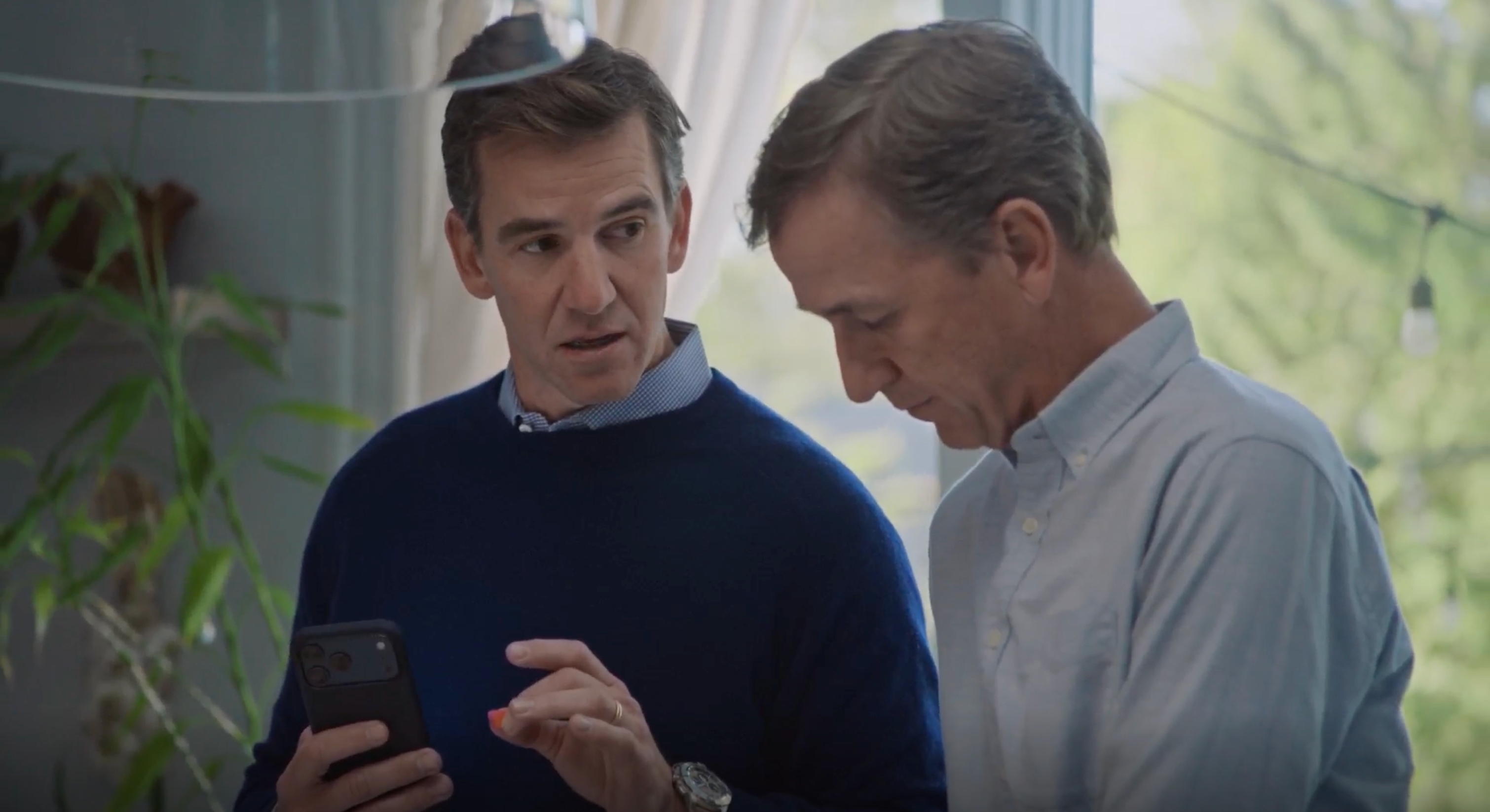 Two men indoors, one holding a smartphone showing it to the other.