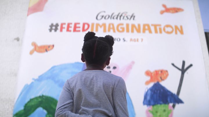 A young girl looking at a Goldfish 'Feed Your Imagination' billboard featuring children’s art. 