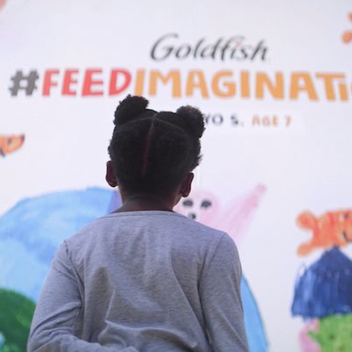 A young girl looking at a Goldfish 'Feed Your Imagination' billboard featuring children’s art.
