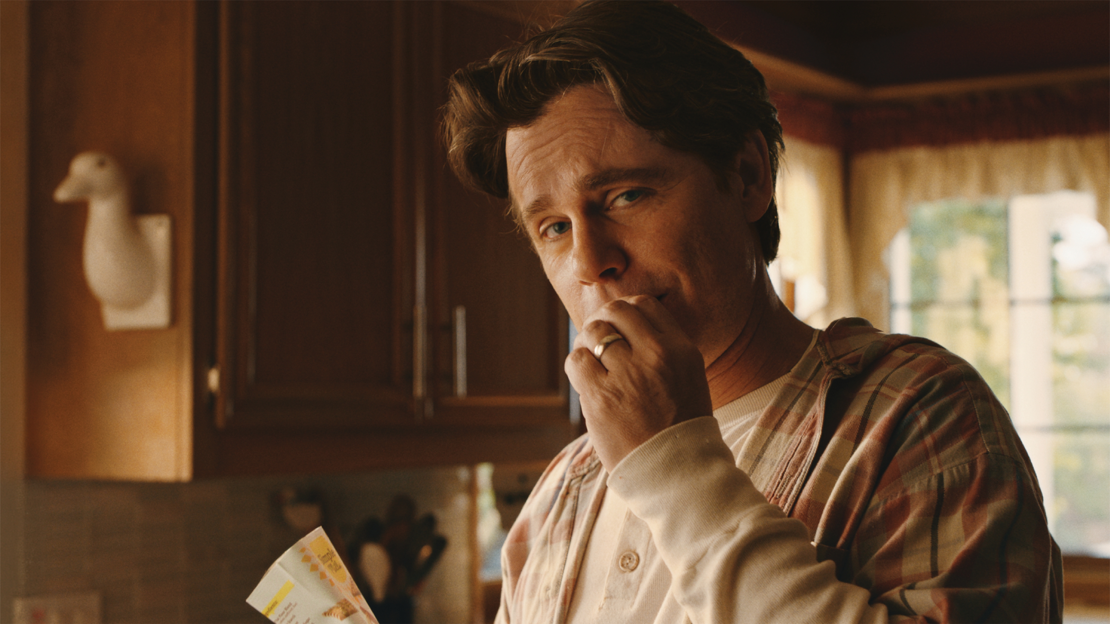 A man stands in a kitchen holding papers and touching his lips, looking toward the camera, with sunlight coming through a window in the background.