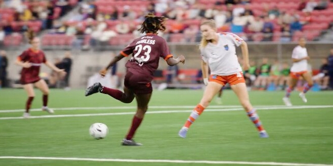Soccer player in maroon jersey dribbles the ball towards a defender in a white and orange kit on a green field. Intense action and focused crowd.