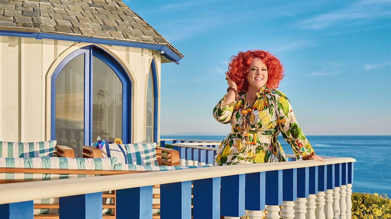 Melissa McCarthy stands on a balcony overlooking a stunning ocean view, smiling as she touches her hair. 