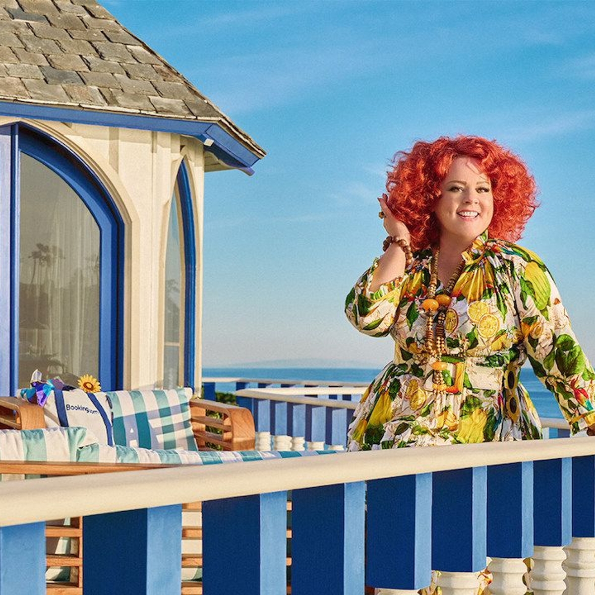 Melissa McCarthy stands on a balcony overlooking a stunning ocean view, smiling as she touches her hair.