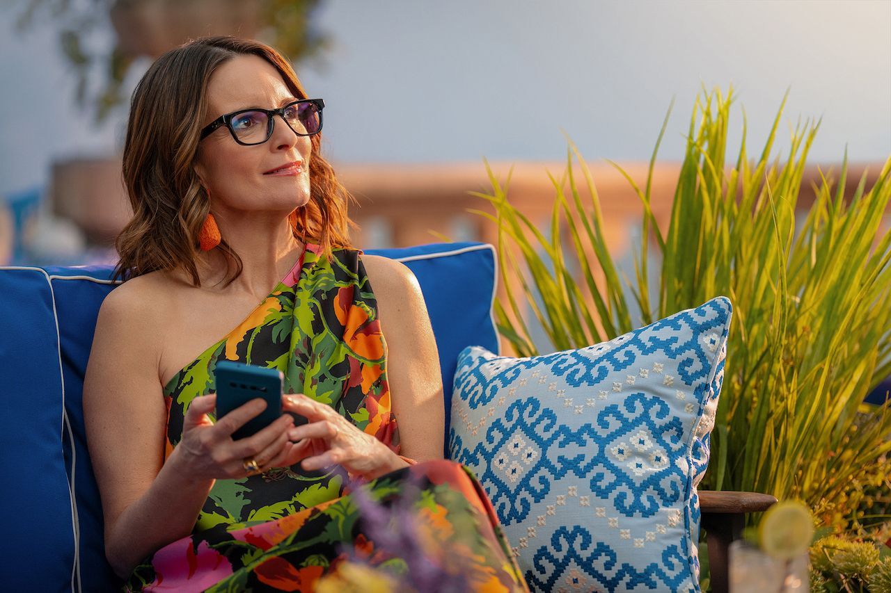 Tina Fey sitting in a soft outdoor chair while holding a phone in her hand. 