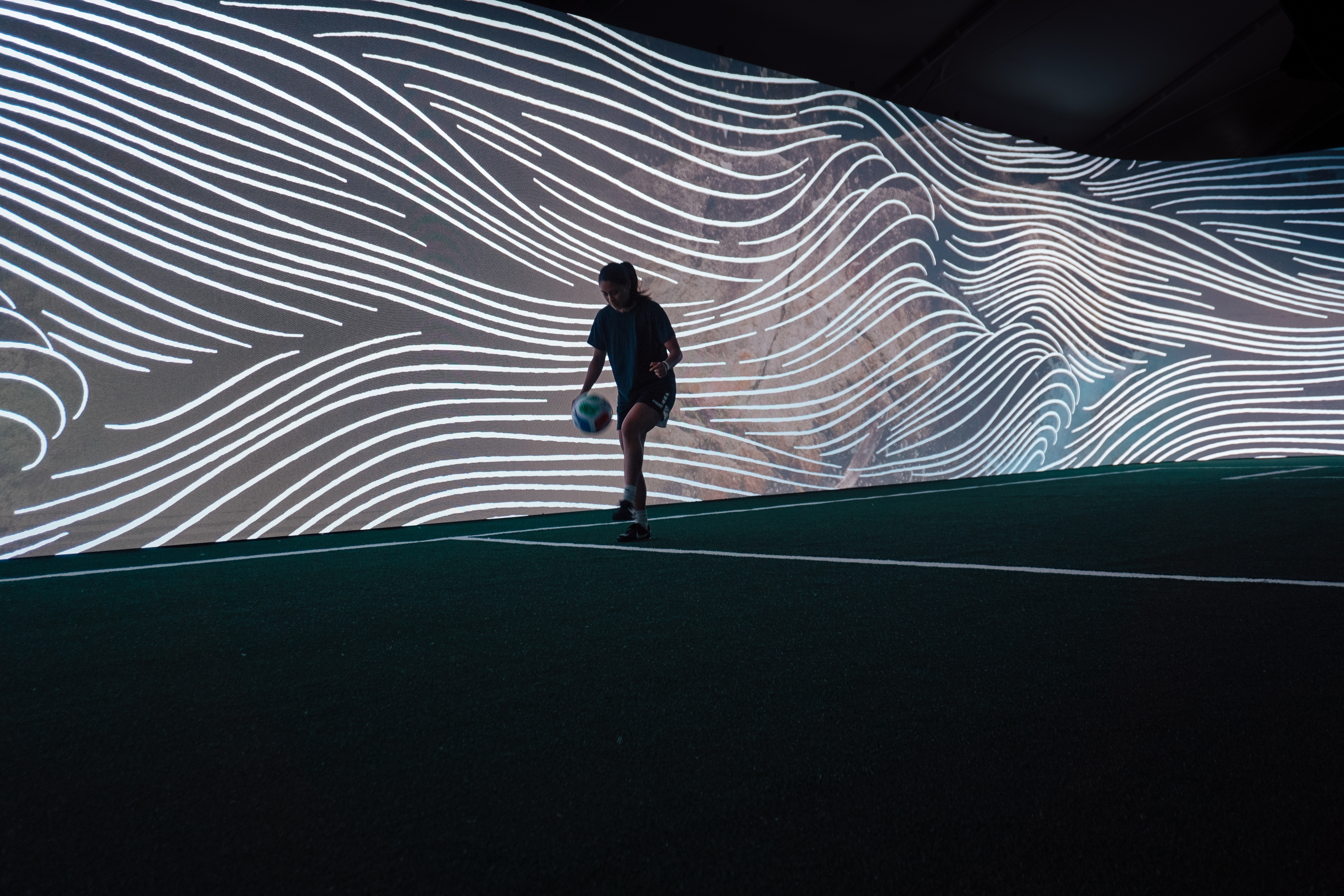 Silhouetted person walks across a dark floor beneath a sweeping digital projection of flowing white lines, creating a futuristic, immersive mood.
