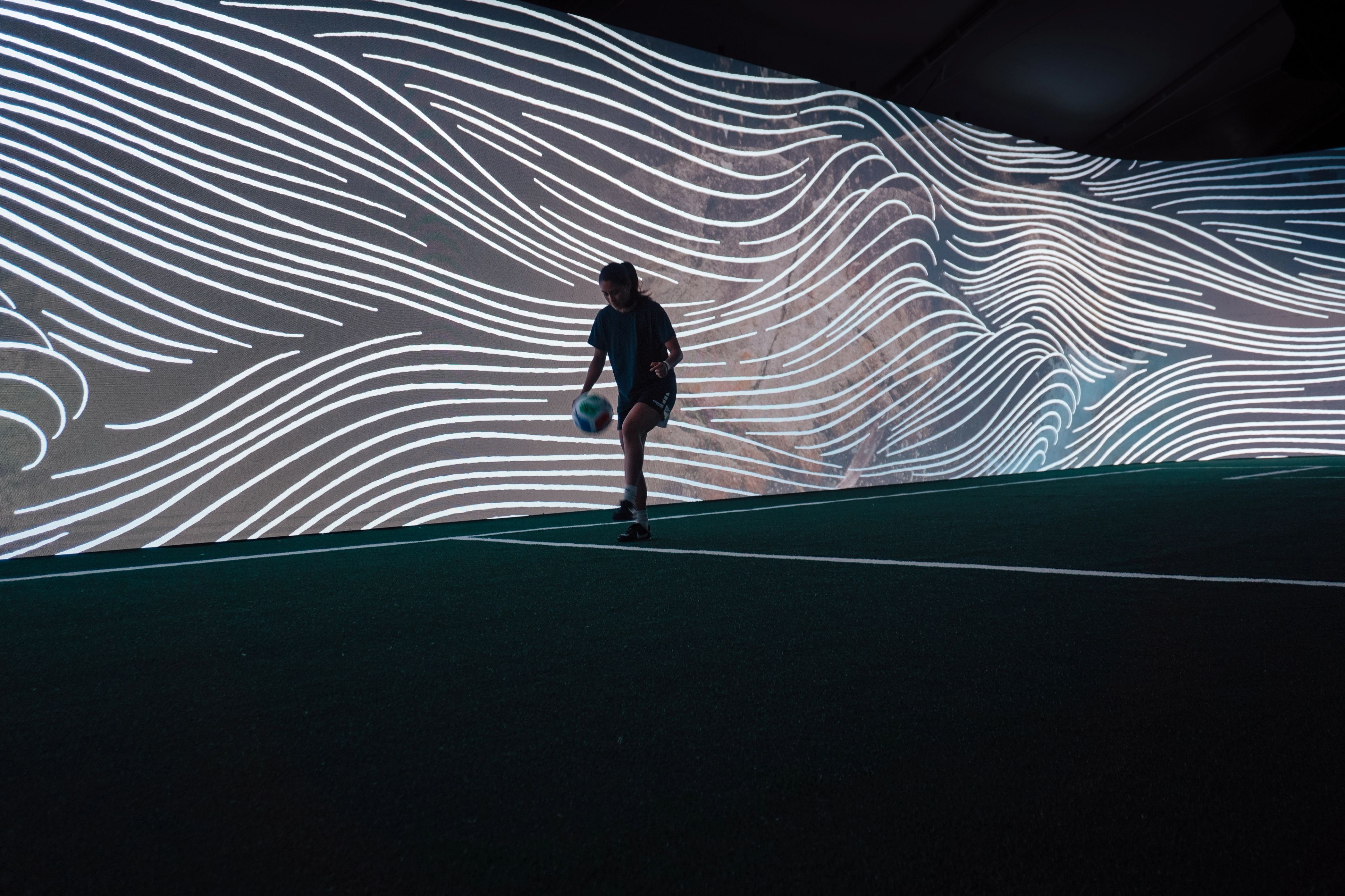 Silhouetted person walks across a dark floor beneath a sweeping digital projection of flowing white lines, creating a futuristic, immersive mood.